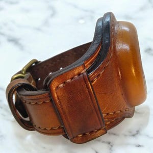 Puede incluir: Una correa de reloj de cuero marrón hecha a mano con cierre de hebilla. La correa presenta costuras detalladas y una apariencia rica y texturizada. La correa del reloj está diseñada para sujetar un reloj de forma segura.