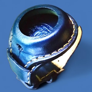 Pode incluir: Um item de couro azul escuro feito à mão, com uma abertura circular e um design em camadas. As bordas são costuradas com linha amarela, e há uma peça de destaque amarelo claro. O objeto é provavelmente um pequeno recipiente ou suporte.