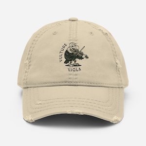 May include: A beige baseball cap with a distressed finish, displaying a cartoon vulture playing a violin. The words "VULTURE" and "VIOLA" are printed above and below the image. The cap has a worn appearance.
