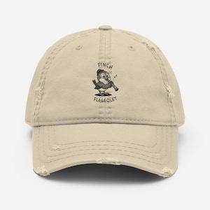 Könnte beinhalten: Abgenutzte beige Baseballkappe mit einem Cartoon-Fink, der eine Flageolet spielt. Der Fink ist schwarz-weiß, trägt einen Hut, und der Text "FINCH FLAGEOLET" steht über und unter dem Vogel. Die Kappe hat einen gebogenen Schirm.