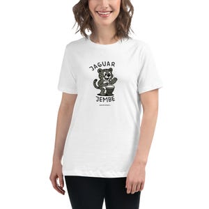 May include: White t-shirt featuring a cartoon jaguar playing a djembe drum. The text "JAGUAR JEMBE" is arched above the jaguar illustration in black. The shirt is a relaxed fit, suitable for casual wear.
