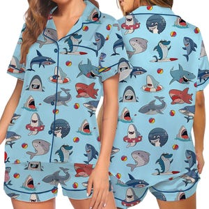 May include: Light blue pajama set featuring a playful shark pattern. The short-sleeved top and shorts are covered in various shark illustrations, beach balls, and surfboards. The set includes a collared shirt with buttons and matching shorts.