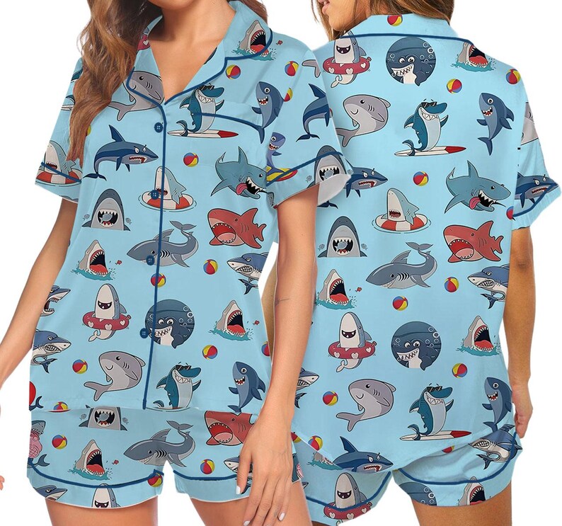 May include: Light blue pajama set featuring a playful shark pattern. The short-sleeved top and shorts are covered in various shark illustrations, beach balls, and surfboards. The set includes a collared shirt with buttons and matching shorts.
