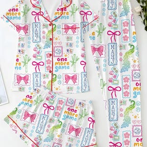 May include: A white pajama set featuring a short-sleeved button-down top, shorts, and long pants. The fabric has a colorful print of pink bows, green bamboo, dice, peacocks, and the text "one more game" and "MAHJ" in blue and red.