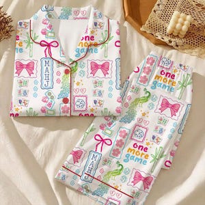 May include: A white pajama set featuring a colorful pattern of mahjong tiles, pink bows, green bamboo stalks, peacock feathers, and dice. The top has a collared neckline with red piping and red buttons. The text "one more game" and "MAHJ" is printed.
