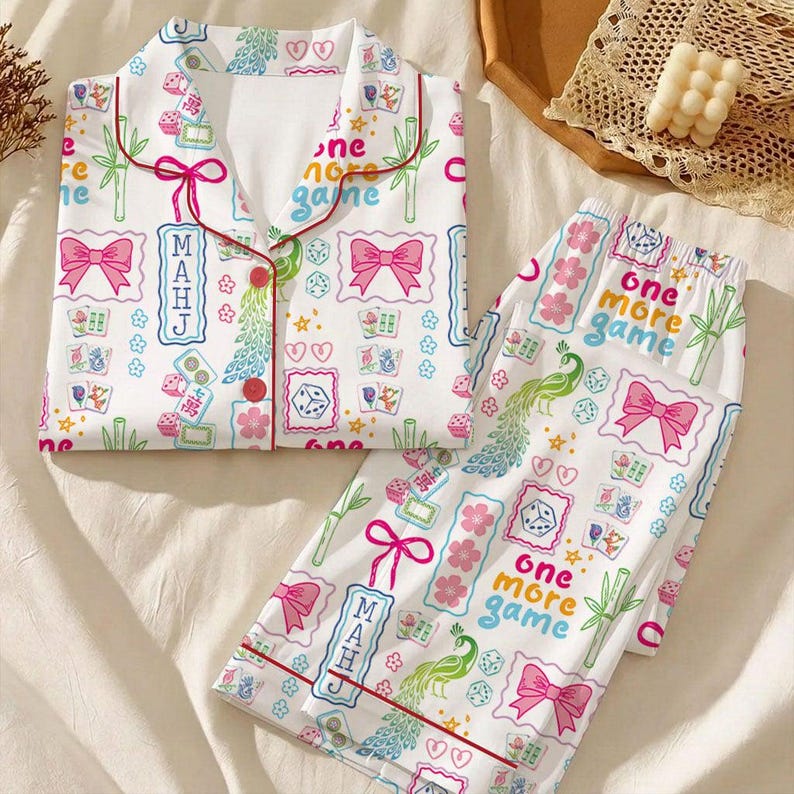May include: A white pajama set featuring a colorful pattern of mahjong tiles, pink bows, green bamboo stalks, peacock feathers, and dice. The top has a collared neckline with red piping and red buttons. The text "one more game" and "MAHJ" is printed.