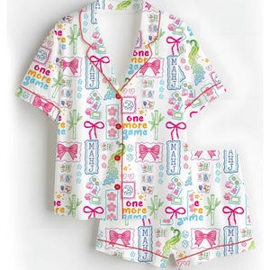 May include: A white short-sleeve pajama set with red piping, featuring a repeating pattern of pink bows, green bamboo, peacocks, dice, and mahjong tiles. The text "one more game" and "MAHJ" is printed throughout the shirt and shorts in various colors.