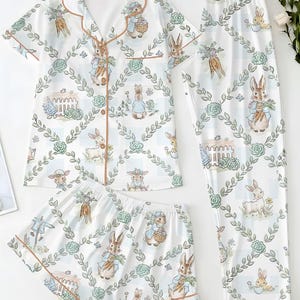 Puede incluir: Conjunto de pijama blanco con estampado de conejitos. El conjunto incluye una camisa de manga corta con botones, pantalones cortos y pantalones largos. El estampado de conejitos presenta conejos, zanahorias y elementos florales. El ribete es de color marr&oacute;n claro.