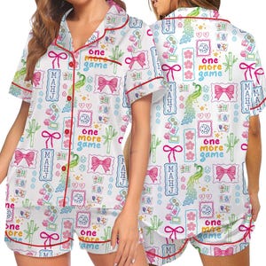 May include: A white pajama set with short sleeves and shorts featuring a red piped trim. The fabric has a colorful pattern of pink bows, green bamboo stalks, dice, peacocks, and the text "one more game" and "MAHJ" in blue and pink.