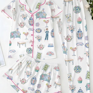 May include: A white pajama set featuring a short-sleeve button-down shirt, shorts, and long pants. The fabric is decorated with a colorful print of teapots, cupcakes, floral arrangements, cocktails, and figures, all accented with bright pink piping.