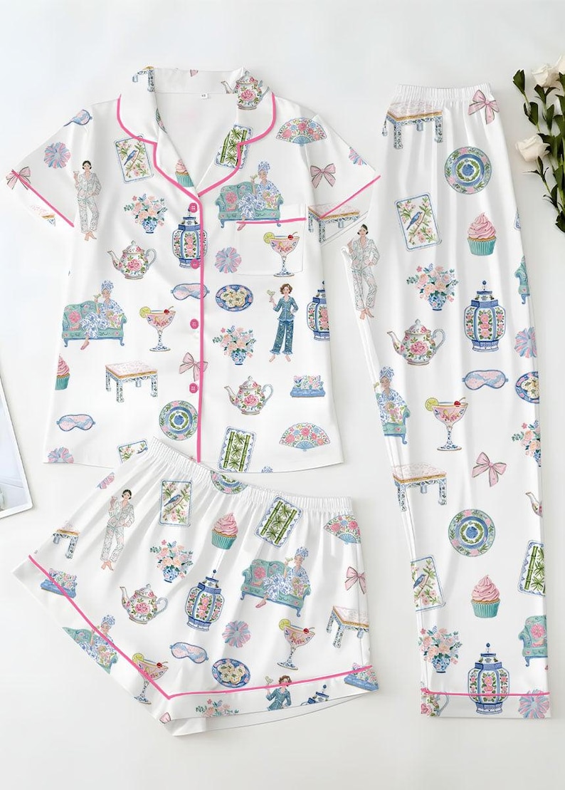May include: A white pajama set featuring a short-sleeve button-down shirt, shorts, and long pants. The fabric is decorated with a colorful print of teapots, cupcakes, floral arrangements, cocktails, and figures, all accented with bright pink piping.