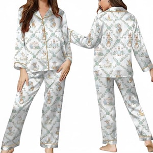 Puede incluir: Conjunto de pijama de sat&eacute;n blanco con un estampado repetido de conejitos, cestas y elementos florales. La parte superior de manga larga y los pantalones tienen ribetes dorados. El pijama tiene una combinaci&oacute;n de colores claros y pastel.