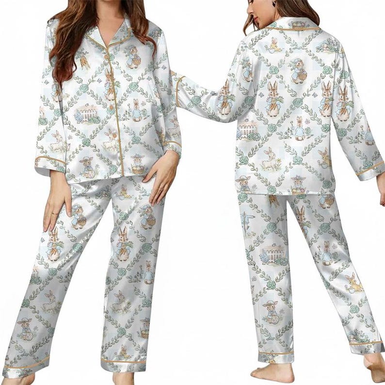 Puede incluir: Conjunto de pijama de sat&eacute;n blanco con un estampado repetido de conejitos, cestas y elementos florales. La parte superior de manga larga y los pantalones tienen ribetes dorados. El pijama tiene una combinaci&oacute;n de colores claros y pastel.