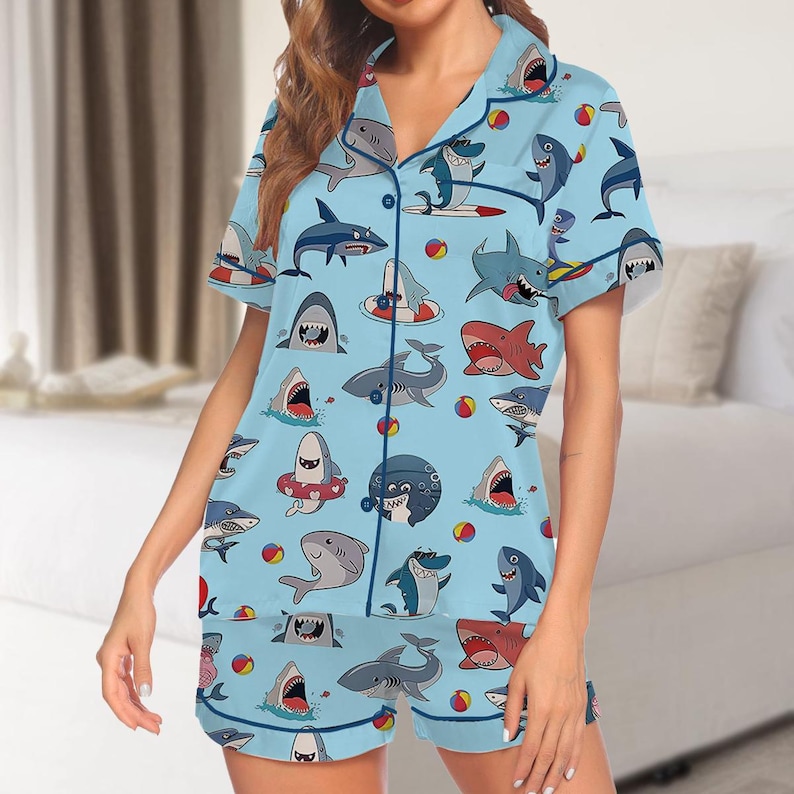 May include: Light blue pajama set featuring a playful shark pattern. The short-sleeved top and shorts are adorned with various shark illustrations, beach balls, and life preservers. The set has navy blue trim and a button-down front.