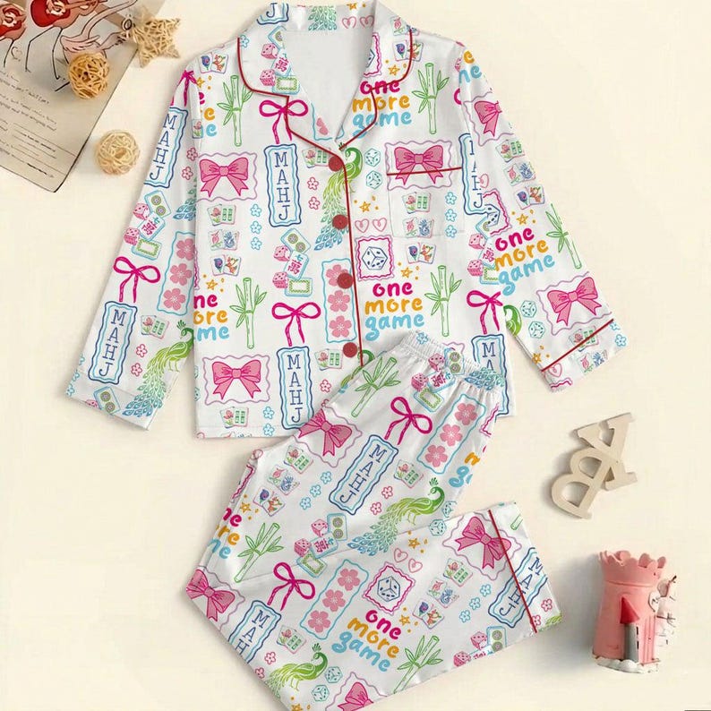 May include: A white long-sleeve pajama set with a button-down top and matching pants. The fabric features a colorful print of pink bows, green bamboo stalks, dice, mahjong tiles, and the text "MAHJ" and "one more game" in blue and red.