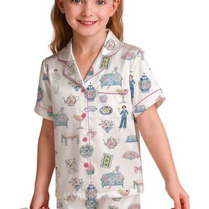 May include: A short-sleeved button-down pajama set in cream satin featuring a colorful repeating print of teapots, floral arrangements, decorative fans, cupcakes, and figures lounging on sofas, finished with pink piping along the edges.