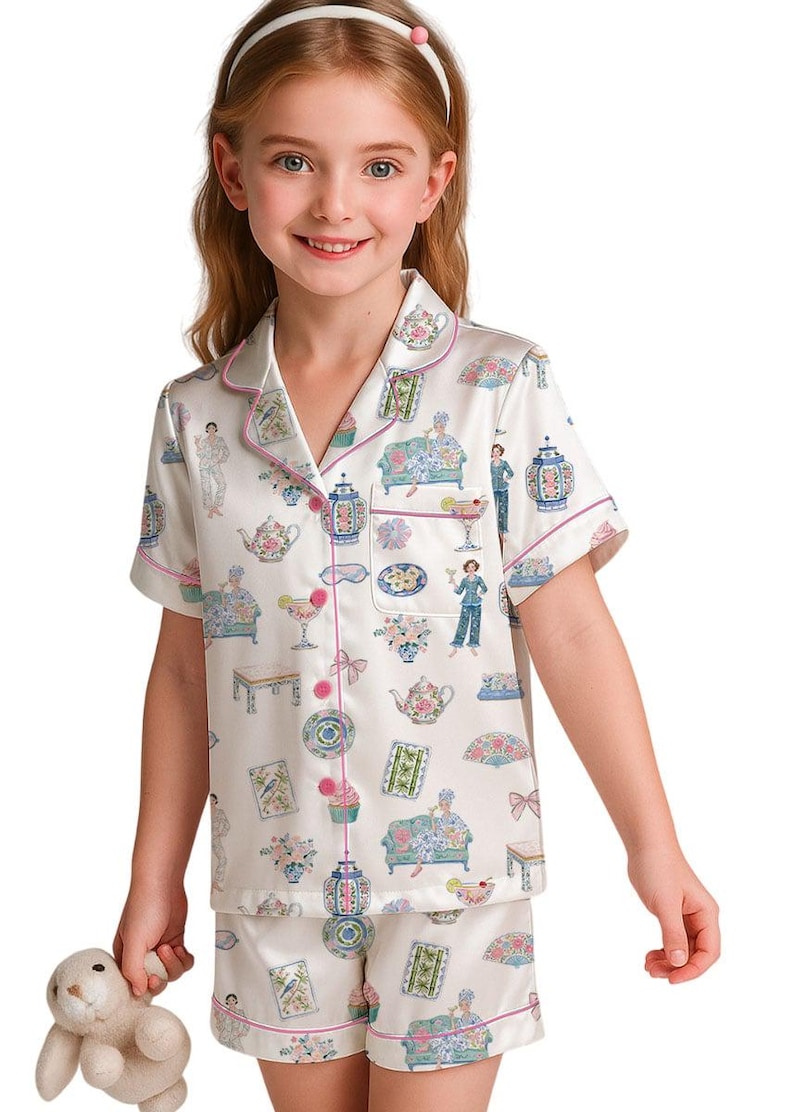 May include: A short-sleeved button-down pajama set in cream satin featuring a colorful repeating print of teapots, floral arrangements, decorative fans, cupcakes, and figures lounging on sofas, finished with pink piping along the edges.