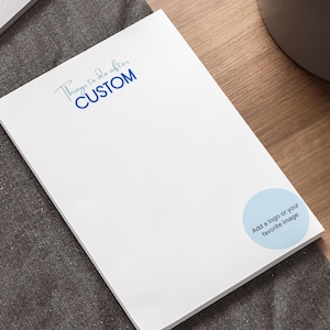 May include: A white notepad with the text "Things to do after CUSTOM" in blue script. A light blue circle with the text "Add a logo or your favourite image" is on the bottom right. The notepad is on a wooden desk.