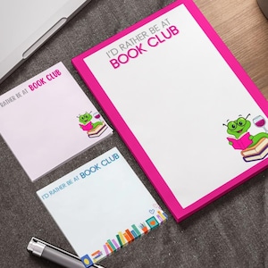 May include: Three white notepads with the text "I'D RATHER BE AT BOOK CLUB" in pink and blue. One notepad has a pink border and a cartoon frog reading a book. The other notepad has a colorful bookshelf design.