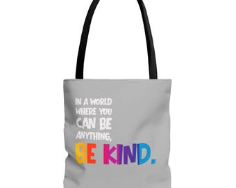 Könnte beinhalten: Eine graue Tragetasche mit schwarzen Griffen. Die Tasche zeigt den Text "IN A WORLD WHERE YOU CAN BE ANYTHING, BE KIND." in Weiß und Regenbogenfarben. Die Tasche ist zum Tragen von Gegenständen konzipiert.