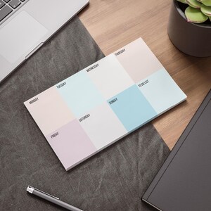 May include: A pastel-colored weekly planner pad with sections for each day of the week and a to-do list. The pad is on a wooden desk next to a laptop, pen, notebook, and a potted plant.