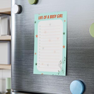 May include: A rectangular notepad with the text "LIFE OF A BUSY GIRL" in red and green. The notepad has a light blue background with star and musical note graphics. It is attached to a silver refrigerator with magnets.