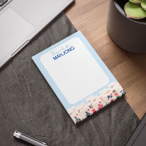 May include: A light blue notepad with the text "Things to do after MAH JONG" in blue. The bottom of the notepad features a design of Mahjong tiles. The notepad is on a wooden desk next to a laptop and a pen.