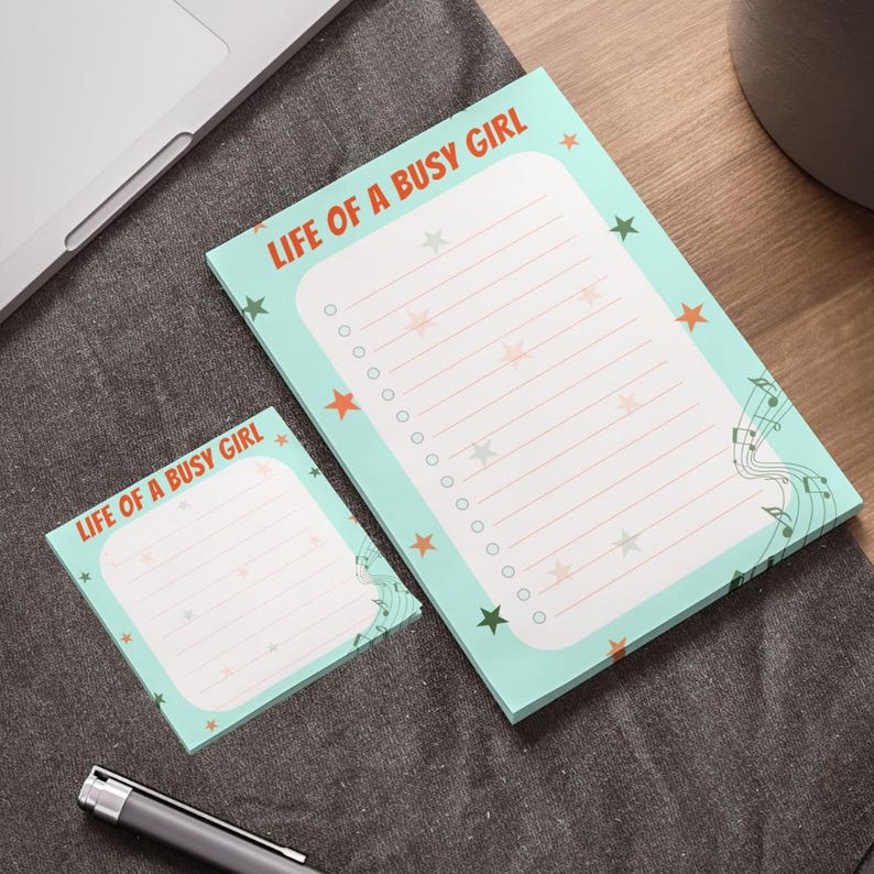 May include: Two notepads with the text "LIFE OF A BUSY GIRL" in orange. The pads are light blue with star and musical note designs. One notepad is smaller than the other. The larger notepad has a list of circles and lines.