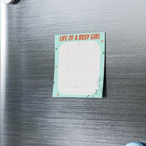May include: A rectangular notepad with the text "LIFE OF A BUSY GIRL" in red and orange. The notepad has a light blue background with white lines for writing, star accents, and musical notes.
