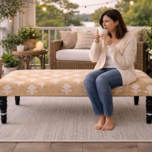 May include: A woman sits on a patterned bench on a porch, holding a white mug. The bench has black legs and a beige floral design. The porch is decorated with plants, candles, and a wicker sofa. A rug is on the floor.