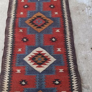Pode incluir: Um tapete corredor retangular com um padrão geométrico. O tapete apresenta um fundo vermelho com formas de diamante azuis e brancas e bordas marrons. O design inclui motivos geométricos repetidos.