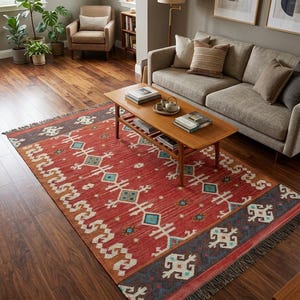 Puede incluir: Una alfombra rectangular con una base de color rojo vibrante y patrones geom&eacute;tricos intrincados en tonos azul, blanco y naranja. La alfombra incluye un borde azul oscuro con motivos blancos y est&aacute; rematada con flecos en los bordes cortos.