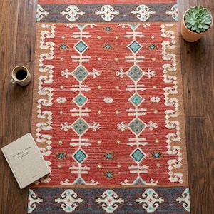 Puede incluir: Una alfombra rectangular con una base de color rojo vibrante y un patr&oacute;n geom&eacute;trico en blanco, turquesa y gris oscuro. La alfombra incluye un motivo de diamantes repetitivo, un borde ancho con s&iacute;mbolos estilizados y flecos grises en los extremos.