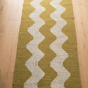 May include: A rectangular woven runner rug featuring a repeating vertical zigzag pattern in mustard yellow and off-white. The rug has a textured fabric construction and is finished with short, dark gray fringe tassels along both narrow ends.
