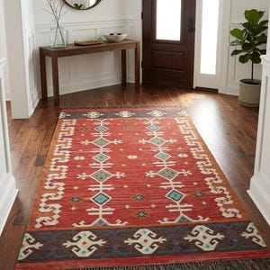 Puede incluir: Una alfombra rectangular con una base de color rojo vibrante y patrones geom&eacute;tricos intrincados en turquesa, blanco y naranja. La alfombra incluye un borde azul oscuro con motivos blancos repetitivos y flecos oscuros en los bordes cortos.