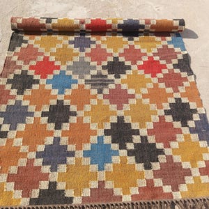 May include: A rolled-up rug with a geometric pattern in shades of red, orange, yellow, blue, and black. The rug has a repeating diamond design. It has a fringe along the bottom edge.