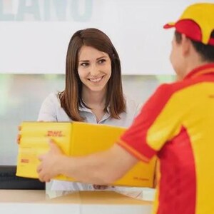 Puede incluir: Una caja de env&iacute;o de cart&oacute;n amarilla con el logotipo rojo de DHL impreso en el lateral. La caja es sostenida por una persona que viste una camisa de uniforme amarilla y roja con una gorra a juego, mientras otra persona la recibe.