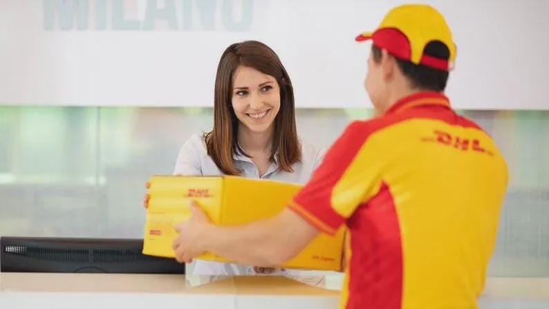 Puede incluir: Una caja de env&iacute;o de cart&oacute;n amarilla con el logotipo rojo de DHL impreso en el lateral. La caja es sostenida por una persona que viste una camisa de uniforme amarilla y roja con una gorra a juego, mientras otra persona la recibe.
