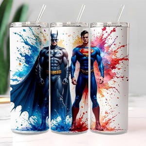 Puede incluir: Tres vasos de acero inoxidable blancos con pajitas transparentes. Cada vaso presenta un dise&ntilde;o de superh&eacute;roe: Batman con un traje y capa azul oscuro, y Superman con un traje azul con el logo "S" rojo. El fondo tiene salpicaduras de pintura coloridas.