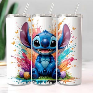 Puede incluir: Vaso blanco con un personaje de dibujos animados azul con grandes orejas y una amplia sonrisa, sobre un fondo de salpicaduras de pintura coloridas. El diseño incluye mariposas y huevos de Pascua. El vaso tiene una pajita transparente y una tapa plateada.