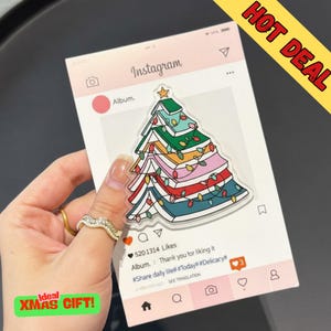 Może przedstawiać: Przezroczysta akrylowa ozdoba choinkowa z kolorowym wzorem, przypominającym stos książek, ozdobiona światełkami i gwiazdką. Ozdoba umieszczona jest na różowym tle w stylu Instagram z tekstem "Instagram" i "Album."