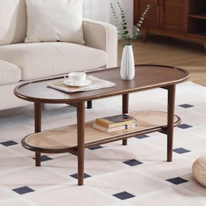 May include: Dark brown oval coffee table with a woven rattan shelf. The table has a curved design and is supported by four legs. A white vase, a cup and saucer, and books are on the table. The table is on a patterned rug.