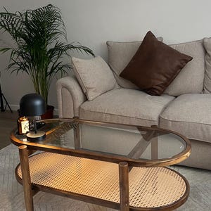 May include: An oval coffee table with a glass top and a lower shelf made of woven rattan. The table has a wooden frame and is placed on a white rug. A black lamp and a candle sit on the table. A beige sofa and a brown leather cushion are in the background.