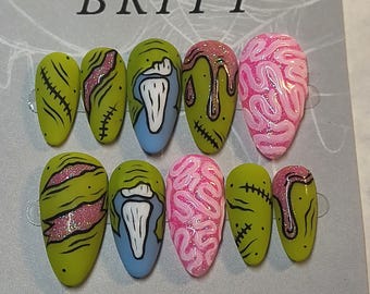 May include: A set of ten almond-shaped press-on nails featuring a zombie-inspired theme with matte olive green, light blue, and pink brain textures. Designs include black stitched scars, pink glittery wounds, and dripping slime accents on a display card.