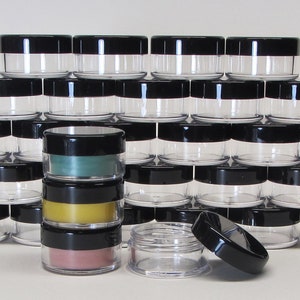 May include: A stack of clear plastic containers with black lids. The containers are stacked in a pyramid shape. There are three containers in the front that are filled with different colored substances. The containers are approximately 1.5 inches in diameter.