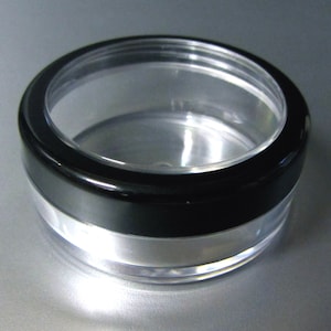 May include: A clear plastic round container with a black lid. The container is designed for storing cosmetics or other small items.