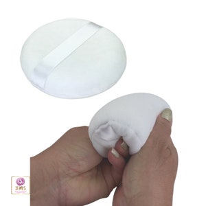 May include: A white round makeup puff with a white satin ribbon. The puff is being squeezed in a hand.