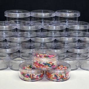 May include: A stack of clear, round plastic jars with screw-on lids. Some jars are filled with colorful sprinkles. The jars are arranged in a pyramid shape, with several jars in the foreground and many more stacked in the background.