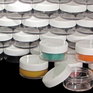 May include: A collection of small, clear plastic jars with white screw-on lids. Some jars are filled with colorful contents, including yellow, orange, and teal. The jars are stacked, with one lid removed, showcasing the product.