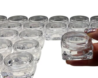 May include: A group of clear plastic jars with lids. The jars are square shaped and have a textured surface. The jars are arranged in a staggered pattern. One jar is being held by a hand.